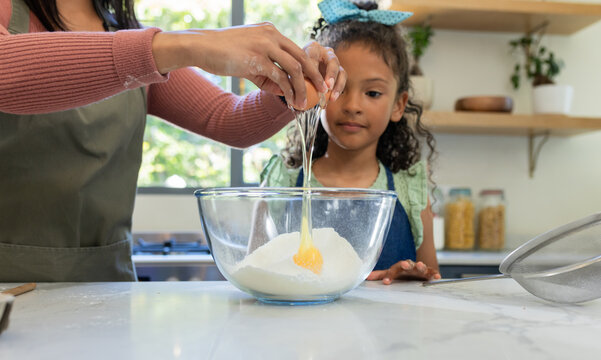 Mother and daughter wearing aprons cracking egg into bowl with sugar at kitchen counter in sunlight