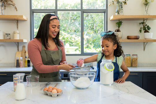 Mother and daughter measuring flour with cup and sifting with sieve at marble countertop in kitchen
