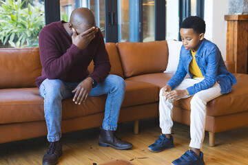 African American father and son sitting on brown leather sofa in living room, expressing distress