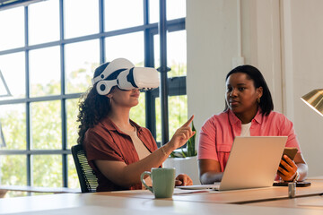 African American female coworkers testing VR headset while noting feedback on laptop near window