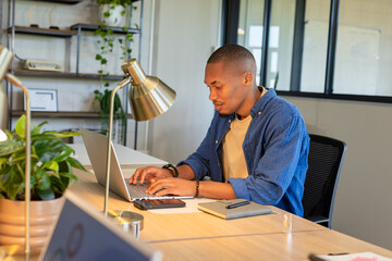 African American man typing on laptop at modern office workspace beside desk lamp and smartphone