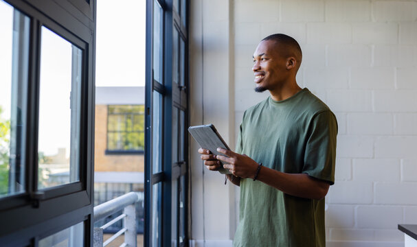 African American man standing in office holding tablet gazing through black windows, copy space