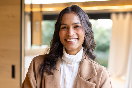Woman wearing white turtleneck and beige coat while smiling in lounge by window showing foliage