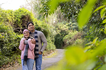Diverse couple embracing while walking along trail through foliage wearing knit scarves, copy space
