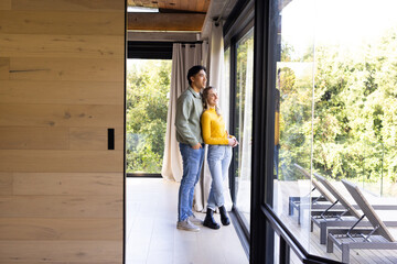 Diverse couple standing by sliding glass door framed by curtains, overlooking terrace lounge chairs