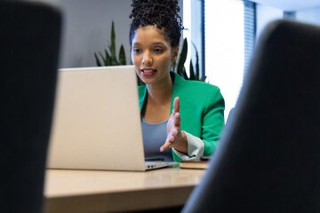 African American woman in green blazer speaking while gesturing at conference table with laptop