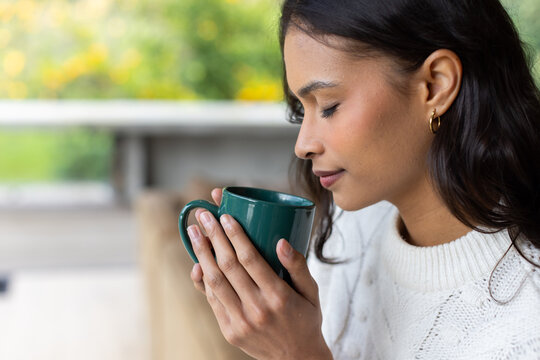 Woman sitting on wooden patio bench holding green ceramic mug and inhaling drink aroma, copy space