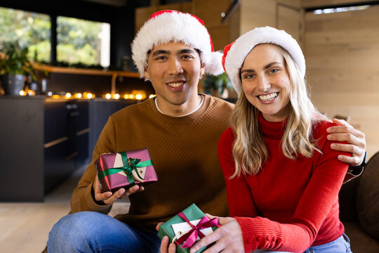 Diverse couple sitting on sofa in cozy living room wearing Santa hats and holding gift boxes - Powered by Adobe