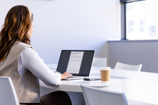 Office worker typing on slim laptop at white conference table with coffee cup, smartphone, chairs