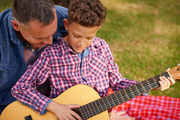 Playing, guitar and father with child in nature for learning, bonding or musical hobby together. Happy, love and dad teaching boy kid song with acoustic string instrument on picnic outdoor in park.
