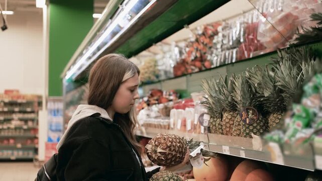 A girl in a black jacket with long hair chooses pineapples in a hypermarket.