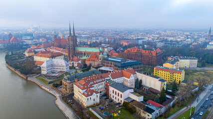 Obraz premium Aerial view of Wrocław’s Ostrów Tumski with the Oder, cathedral, historic buildings, red rooftops and the city skyline in light fog