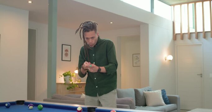 Man with dreadlocks entering home, chalking cue at pool table, aligning shot and striking cue ball