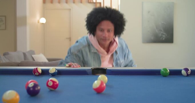 After lining up shot, African American man striking cue ball on pool table at home, celebrating