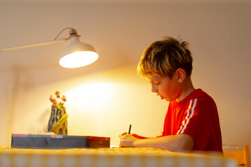 Youth studies diligently, Teenager focusing on schoolwork, Adolescent engaged in writing at neatly arranged desk, Teenager concentrating on homework under cozy lamp surrounded by personal items