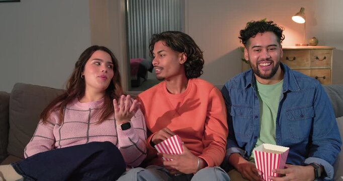 Diverse friends holding popcorn boxes on sofa in living room, glancing, explaining joke, laughing