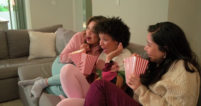 Diverse female friends eating popcorn then reacting to jump scare and laughing on sectional sofa