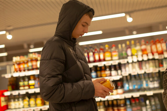 Teen in hooded jacket inspecting label, student researching ingredients for school project, standing in supermarket aisle, holding bottle, shelves stocked with colorful products, warm overhead - Powered by Adobe