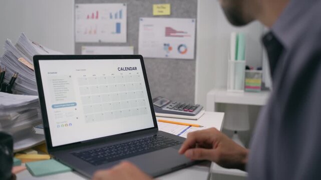 A hand checks a laptop calendar to manage a deadline timeline with an overworked task planner in the office.