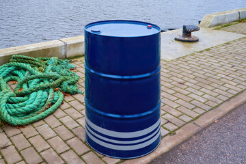 Blue metal barrel standing on a quay near coiled green rope and waterfront pavement, representing industrial storage, marine equipment and harbor infrastructure.