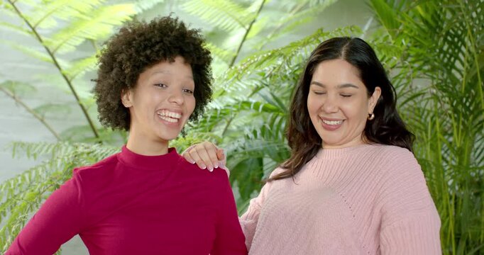 Diverse female friends by brick wall and ferns, turning toward friend, laughing then pouting