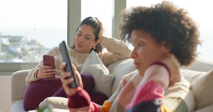 Diverse female friends reclining on home sofa scrolling smartphone and tablet, basking in sunlight