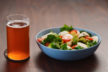Salad with avocado, cherry tomatoes, romaine and mozzarella in blue bowl and glass of kombucha on walnut table