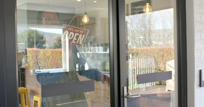 African American man flipping closed sign to open at cafe, opening glass door and welcoming guests