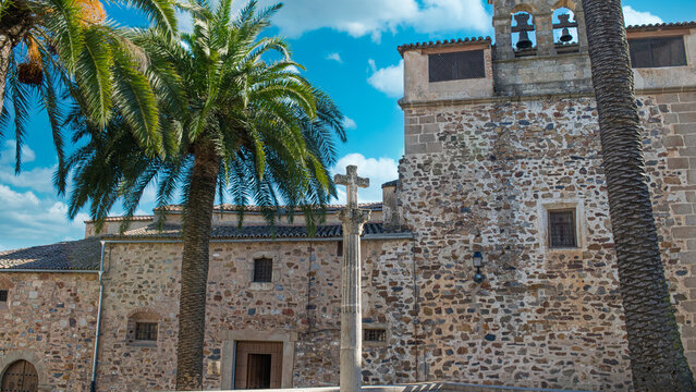 Plaza con palmeras y cruz de piedra con el convento de Santa Clara de fondo en la ciudad de C&aacute;ceres, Espa&ntilde;a