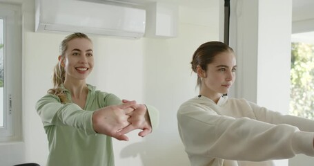 Female duo interlacing fingers, extending wrists in home gym under AC, band strap by foliage window