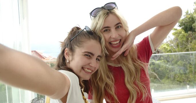 Two female friends holding smartphone for selfie, wearing sunglasses and lip-syncing in sunroom