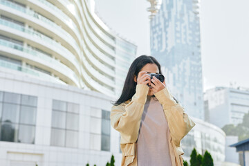 Woman with camera in an urban cityscape, capturing skyline and modern buildings outdoors in daylight, wearing a light jacket, candid street photography moment, travel vibes, urban exploration