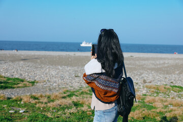 Beach scene of a traveler capturing the ocean view with a smartphone, wearing a backpack and layered clothing, sunny sky, calm waves, and open horizon, travel, photography, outdoors