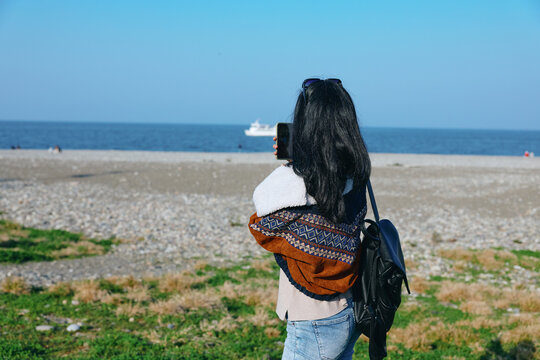 Woman on a sandy beach with backpack gazing at the sea, casual jacket and scarf, exploring coastal landscape and outdoor travel moment - Powered by Adobe