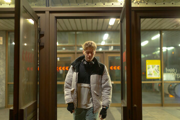 young white man arriving at station in puffer jacket, latenight platform lighting and glass shelter create urban commuter vibe, hands in pockets and steady stride capture candid public transit scene