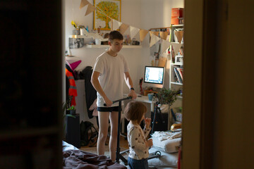 teenagers cluttered bedroom gaming setup, older sibling guiding younger through gameplay and basic coding, warm lamp glow illuminating monitors and posters, tangled cables and scattered clothes,