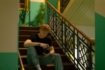 Young man on stairs reviewing camera, student photographer examines LCD screen, adjusting settings, holding DSLR, casual denim and sneakers, smartwatch on wrist, warm teal stairwell light, quiet urban