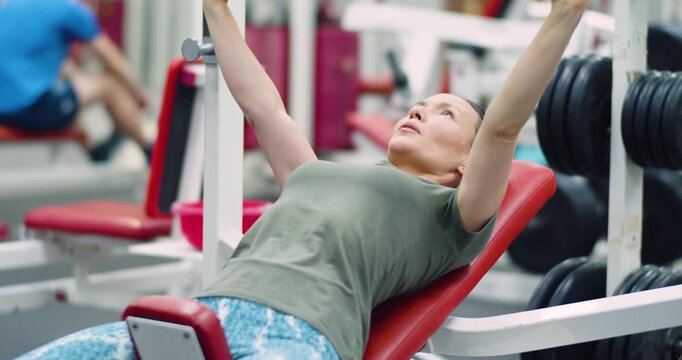 Gym workout. 4k footage of a middle-aged woman performs bench presses in the gym, she trains her chest with bench presses, maintaining proper form and power during her gym workout session.