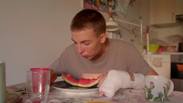 white teen face-first watermelon bite at kitchen table with bandaged hand, juice splatter, concentrated grin,