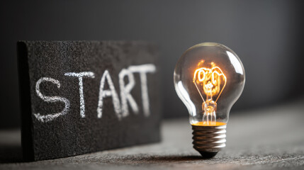 Dramatic low-key close-up of brightly illuminated vintage filament light bulb standing next to small chalkboard slate with the word START written in chalk, symbolizing beginning of new idea or project