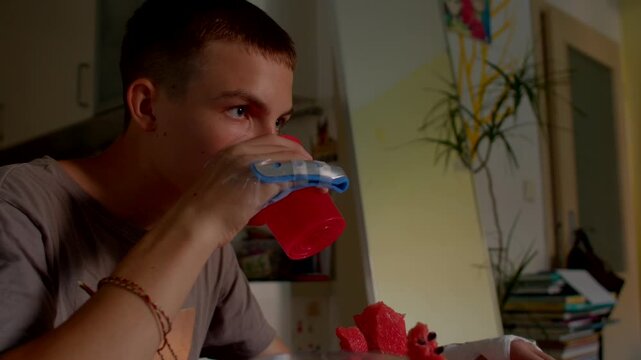 white teen sipping watermelon juice at kitchen table with bandaged hand, red fruit pieces on plate, casual