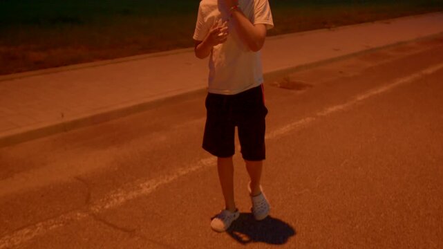 teen boy standing on empty street under orange glow, hands in pockets and relaxed stance, distant city lights