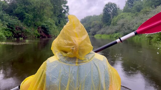 Person in a yellow rain poncho paddling a kayak in rainy weather on the Irbe River, with wet vegetation, calm water and dense forest visible along the riverbanks in Latvia