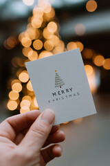Hand shows cheerful seasonal card before illuminated outdoor evergreen with glowing lights.