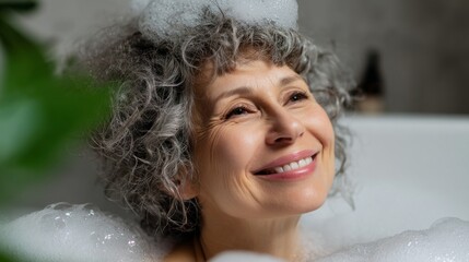 A cheerful senior woman with gray, curly hair smiles brightly while enjoying a luxurious bubble bath. Soft light enhances the peaceful atmosphere, creating a moment of pure relaxation at home