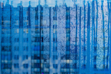 Vertical blue streaks of water and soap suds on a glass window, with a blurred city building facade visible through the pane, creating an abstract urban pattern.
