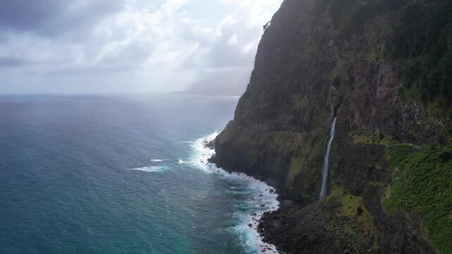 Aerial Madeira coastal scene with a waterfall Veu da Noiva plunging down steep cliffs into the ocean. The rugged landscape merges with the dynamic waves, creating a breathtaking natural spectacle