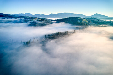 Aerial view of landscape enveloped in thick, rolling clouds. Verdant hills and scattered trees barely peek through, while backdrop of distant mountains completes this serene and dreamlike scene.