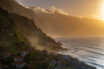 Dramatic aerial view of Madeira's coastline at sunset, showcasing vibrant oranges and blues. The serene ocean, lush mountains, and charming village create a captivating scene