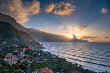 Dramatic aerial view of Madeira's coastline at sunset, showcasing vibrant oranges and blues. The serene ocean, lush mountains, and charming village create a captivating scene